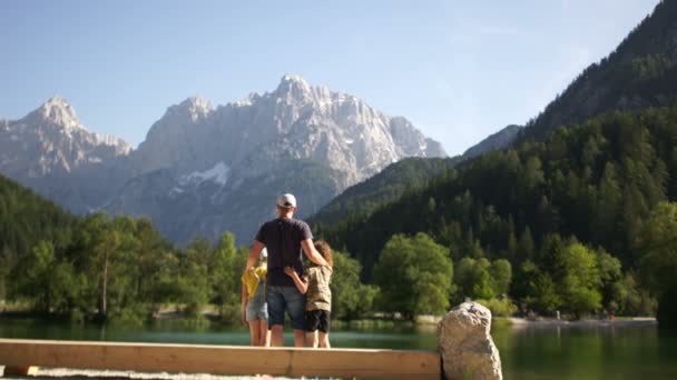 A gyönyörű természeti panorámát csodáló család. Egy apa és a két tizenéves gyermek ül egy padon a parton egy hegyi tó. Nyári szünidő, boldog családi pihenés — Stock videók