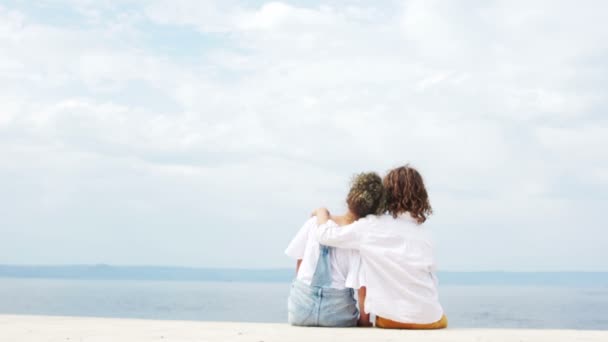 Счастливые дети, мальчик и девочка сидят, обнимаясь на берегу моря, и смотрят вдаль. Летние каникулы — стоковое видео