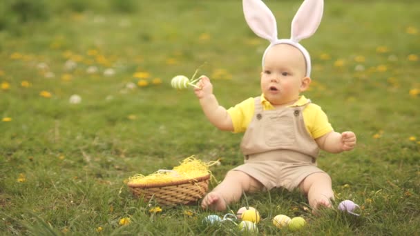 Kleinkind im Anzug des Osterhasen spielt mit Ostereiern, die auf der Wiese im Park sitzen. Frühlingspicknick, glückliche Osterfamilie