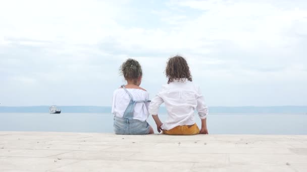 Boy and a girl schoolchildren are sitting next to the seashore and looking into the distance at a sailing ship. Travel concept, summer vacation — Stock Video