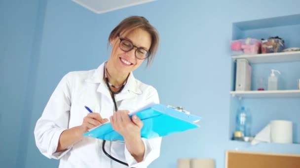 Ženský lékař v bílém lékařském kabátě a s fonendoskop se podívá do rámu a usmívá se s úsměvem s bílými zuby — Stock video