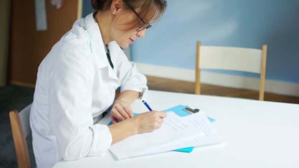 Kvinnlig läkare skriver ett recept fyller ut sjukdomshistoria vid ett skrivbord i ett medicinskt kontor. Internationella läkar dagen — Stockvideo