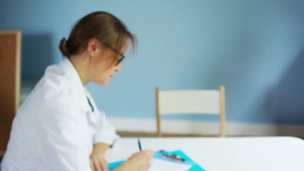 Женщина-врач в белом халате и очках выписывает рецепт на лекарства пациенту, сидящему за столом в медицинском кабинете. Смотрит в камеру и улыбается. — стоковое видео