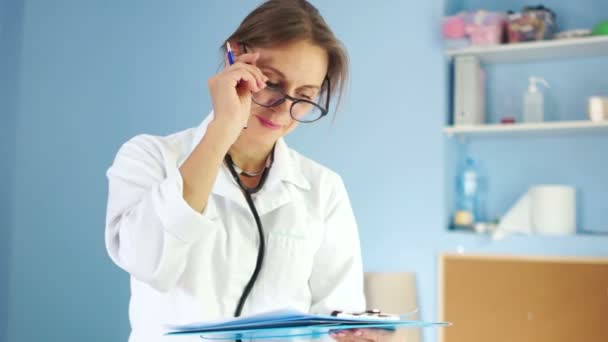 Mulher rigorosa médico com óculos e um casaco branco escreve uma receita em seu escritório. Dia Internacional dos Médicos. Vestido médico — Vídeo de Stock