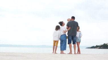 Ebeveynler ve üç çocuk. Babasının kollarındaki çocuk, anne ve babasını öpmek için uzanır ve garip bir şekilde ağzını açar. Büyük bir aile deniz kıyısında dinleniyor.