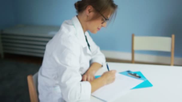 Kinderarzt in weißem Mantel und Brille schreibt einem Patienten, der an einem Schreibtisch in einer Arztpraxis sitzt, ein Rezept für Medikamente. blickt in die Kamera und lächelt — Stockvideo