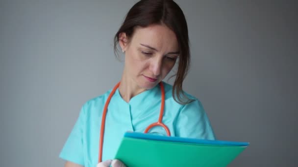 Retrato de una joven doctora mostrando algo o copia de producto o texto de signos, con foder azul, sobre fondo gris. Día Internacional del Médico, medicina y concepto de salud — Vídeos de Stock