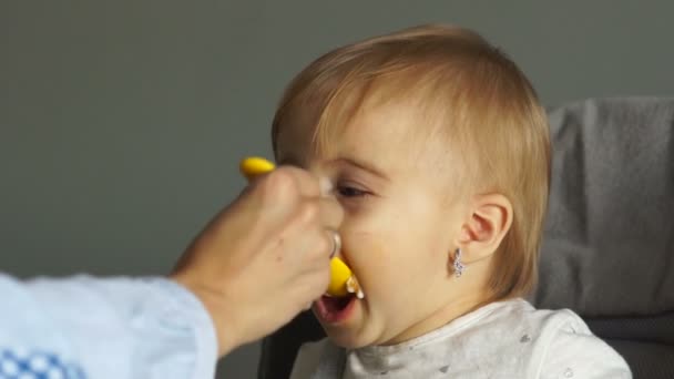 Piękne niebieskookie dziecko siedzi w fotelu i z należytą starannością je puree warzywne, otwierając usta. Mama karmi dziecko łyżką. — Wideo stockowe