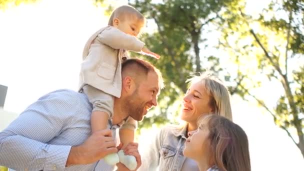 Gelukkige familie met twee kinderen voor een wandeling in het park in de zon. Ouders en twee dochtertjes — Stockvideo