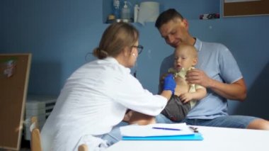 Klinikteki hastaların resepsiyonu. Doktor babasının kucağında oturan bebeği muayene eder. Tıp ve sağlık hizmetleri kavramı