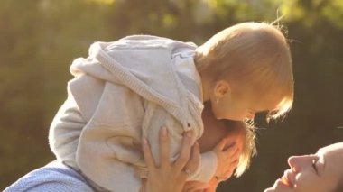 Bir yaşında bir bebek babasının omuzlarında otururken annesi bebeğini öpmek için uzanır. Mutlu bir aile parkta yürüyüşe çıktı.