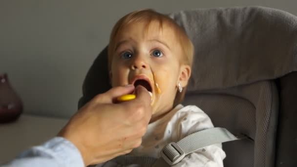 Une mère non identifiée nourrit sa fille d'un an avec une cuillère. L'enfant est assis sur une chaise haute. Manger sainement pour les tout-petits — Video