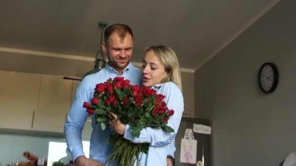 Man gifting bouquet of roses to smiling woman at kitchen. Loving couple with a bouquet of flowers in the kitchen. Newlyweds on Valentines Day cuddle at home — Stock Video