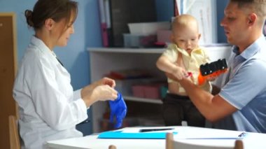 Kadın doktor steteskop kullanarak bebeğin nefes alışını dinliyor. Baba ve bebek çocuk doktoruna geldi.