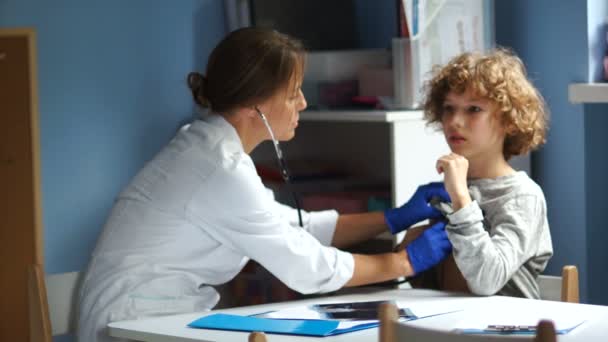 Female doctor listens to the lungs and bronchi in a teenager and writes the result on a medical record. Curly boy teenager at a reception at the pediatrician — ストック動画