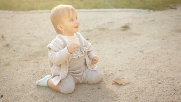 Забавная годовалая девочка, сидящая на песке и играющая с галькой. Девушка в голубом комбинации, счастливое детство — стоковое видео