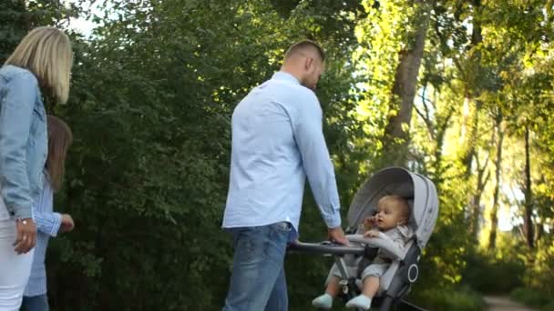 Joven familia feliz caminando en el parque de verano. Padre lleva un cochecito con un bebé, mamá va de la mano con su hija. Vista trasera — Vídeos de Stock