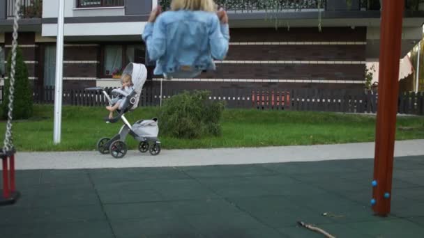 Moederdag, jonge moeder. Een eenzame jonge vrouw zwaait op een speelplaats op de achtergrond van een wandelwagen met haar baby — Stockvideo