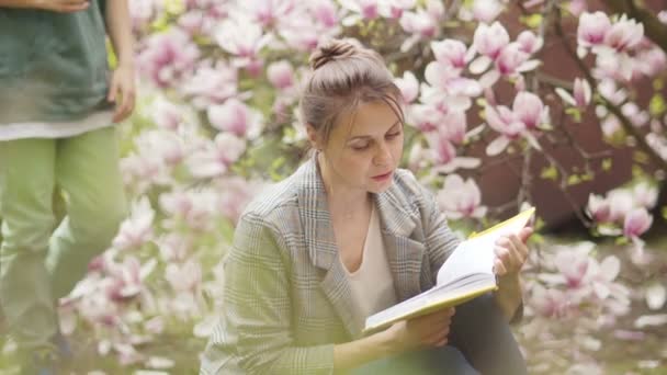 Женщина читает книгу на фоне куста, цветущего розовой магнолией. Кудрявый сын подходит к ней и дает одуванчик. День матери — стоковое видео