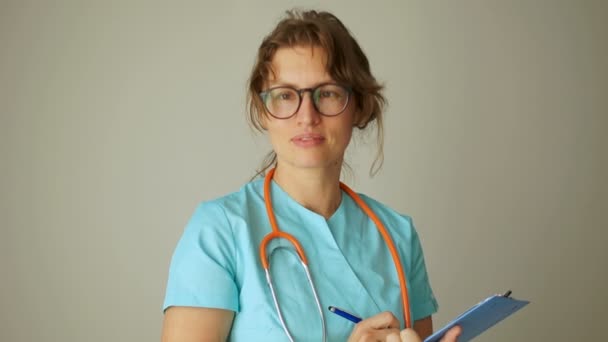 Belle femme médecin dans un manteau médical avec un stéthoscope sur le cou et un dossier dans ses mains regardant la caméra et souriant. Fermer Portrait, Journée internationale des médecins — Video