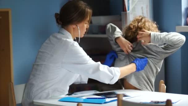 A gyerekorvos megvizsgál egy diákot a fogadáson. Egy sztetoszkóppal hallgatja a tüdőt hátulról. — Stock videók