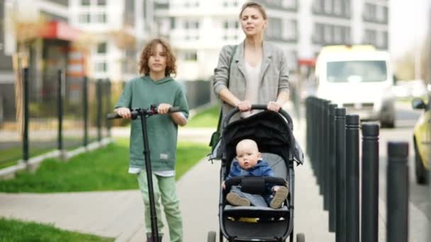 Piękna młoda kobieta z wózkiem spacerowym z synami na ulicy miasta. Zboczony uczeń jeździ na skuterze elektrycznym. Dzień Matki — Wideo stockowe