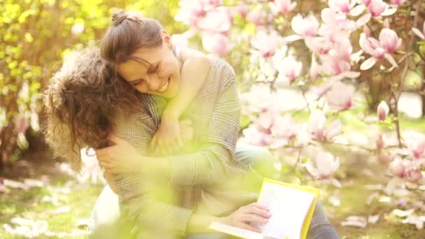 Glücklicher Teenager umarmt seine schöne junge Mutter. Frühlingsporträt auf einem Hintergrund blühender rosa Magnolie. Muttertag — Stockvideo