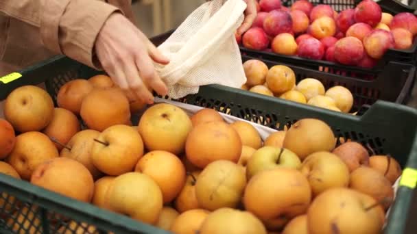 Close plan, hands of a woman in a raincoat buying apples in a supermarket or a vegetable shop. Eco packaging, reusable pads, fabric shopping bags — Stock video