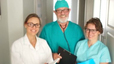 Tıbbi önlüklü ve gözlüklü üç doktor kameraya bakıp gülümsüyor. Uluslararası Doktorlar Günü