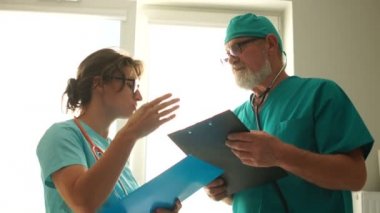 İki doktor hastanenin penceresinin önünde konuşuyorlar. Doktor erkek ve bayan asistan hasta tedavisi için randevuları tartışıyorlar