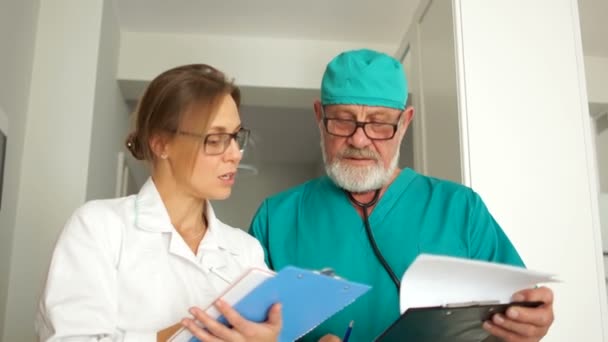 Staff of the medical clinic. Close portrait of two doctors, a mature man and a young woman. Doctors look at the camera and smile. The man has a neat beard and a stethoscope — Stock Video