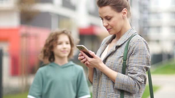 Ώριμη επιχειρηματίας σε ένα δρόμο της πόλης κοιτάζοντας ένα smartphone. Έρχεται ο έφηβος σγουρός γιος της και αγκαλιάζονται χαρούμενα. Ημέρα μητέρων — Αρχείο Βίντεο