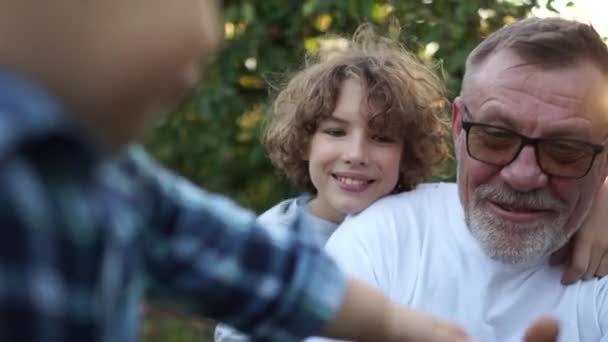 Farfar och hans barnbarn skrattar glatt i trädgården vid solnedgången. Semester i byn, lycklig familj, barn äter ett äpple — Stockvideo