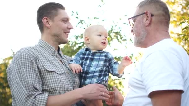 Три поколения одной семьи веселятся в яблоневом саду на закате. Дедушка, сын и внук смеются, дед целует внука — стоковое видео