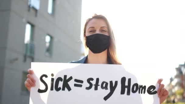 Sjuk - stanna hemma frisk - gå till jobbet. Europeisk protest mot stränga inlåsningsåtgärder under karantänen av coronavirus covid-19. Flicka i mask med affisch står nära staketet i stadshuset — Stockvideo