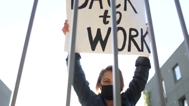 Dziewczyna w czarnej masce z plakatem z powrotem do pracy protesty w pobliżu ogrodzenia organizacji państwowej. Protest społeczny przeciwko środkom ograniczającym podczas kwarantanny covid-19 — Wideo stockowe