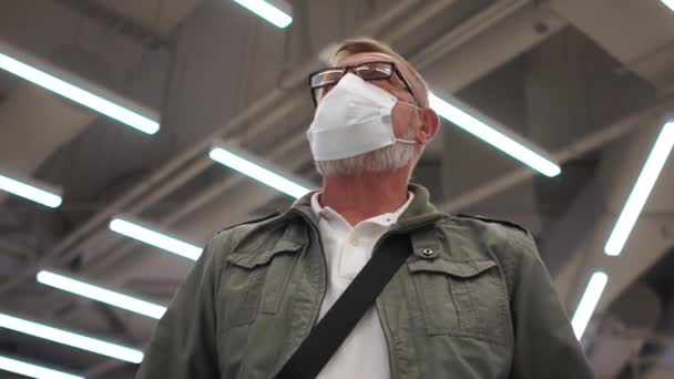 Porträtt av en pensionär i mask, glasögon mot taket med lysrör. Efter karantänlivet, socialt avstånd. Porträtt av en gammal man som bär ansiktsmask vid utbrott av coronavirus — Stockvideo