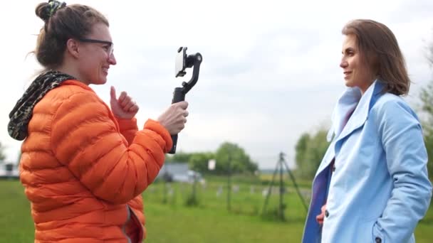 Modell und Bediener mit Smartphone und Stabilisator. Erstellen von Inhalten für einen Videoblog. Zwei Frauen drehen Videos im Park. Daumen hoch wie Geste — Stockvideo