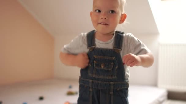 Un niño encantador con un mono de mezclilla está bailando en casa. Feliz infancia. — Vídeos de Stock