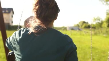 Genç anne salıncaktan inip oğlunu salıncakta sallamaya başladı. Güneşli bir günde anne ve oğul, mutlu çocukluk, anneler günü.