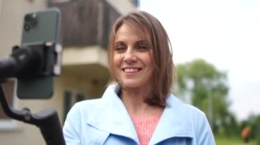 Akıllı telefon kamerasında poz veren orta yaşlı güzel bir kadın. Video blogunda açık hava portresi. Cep telefonu kamera sabitleyici açık hava video blogu kaydeden bir Vlog yazarı