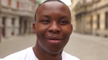 Siyahi adamın portresi çok mutlu, kentin arka planında gülümsüyor. Afro-Amerikalı bir öğrenci şehrin ortasında dururken samimiyetle gülümsüyor. Adam beyaz bir tişört giyiyor.