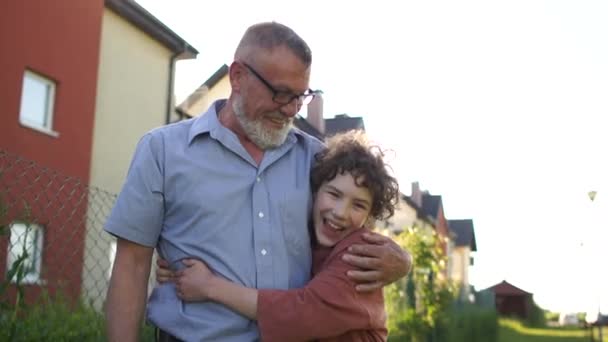 Gelukkige kleinzoon en grootvader knuffelend tegen de achtergrond van een huis in het dorp. Zonsondergang, zomervakantie, vaderdag — Stockvideo