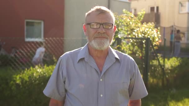 Feliz pensionista con una barba gris y gafas se encuentra cerca de su casa. Sol retrato llamarada, concepto de bienes raíces — Vídeos de Stock