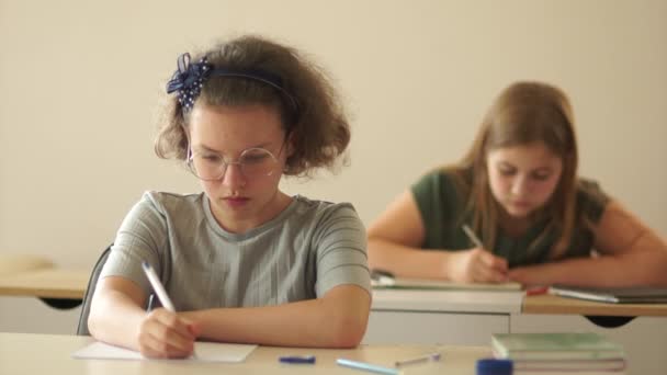 Близкий портрет школьниц-подростков. Они сидят за партами в классе и пишут в блокнотах. Диктация класса — стоковое видео