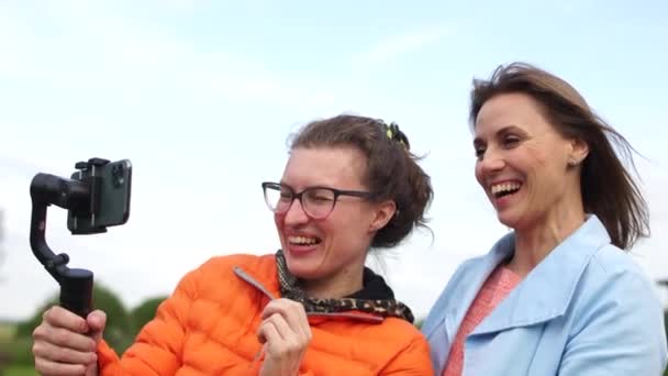 博客的录像。两个女人，一个模特和一个摄影师在镜头前笑着挥手。操作员拿着一部智能手机和一个稳定器 — 图库视频影像