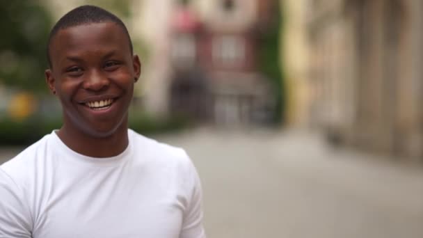 カメラを見ながら笑顔の黒人男性。白いTシャツの若いアフリカ系アメリカ人男性の都市屋外の肖像画 — ストック動画