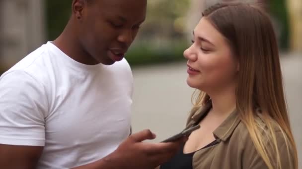 Siyah adam ve beyaz kadın akıllı telefona bakıp neşeyle gülüyorlar. Irklar arası çift şehir sokaklarında eğleniyor. Genç yaşam tarzı konsepti — Stok video