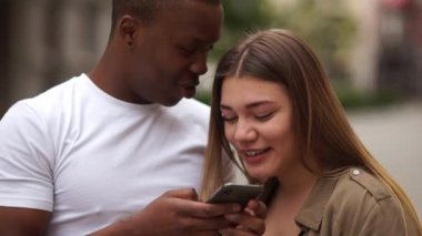 Bir çift melez öğrencinin açık hava portresi. Afrikalı bir adam ve beyaz bir kız akıllı telefona bakıp gülüyorlar.