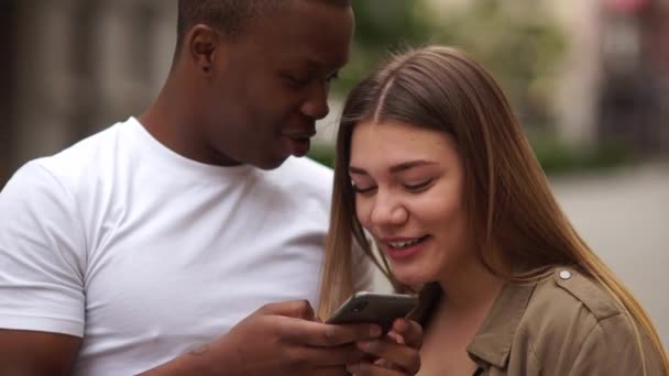 Zewnętrzny portret mieszanej pary studentów. Afryki facet i kaukaski dziewczyna patrzeć na smartfon i śmiać — Wideo stockowe